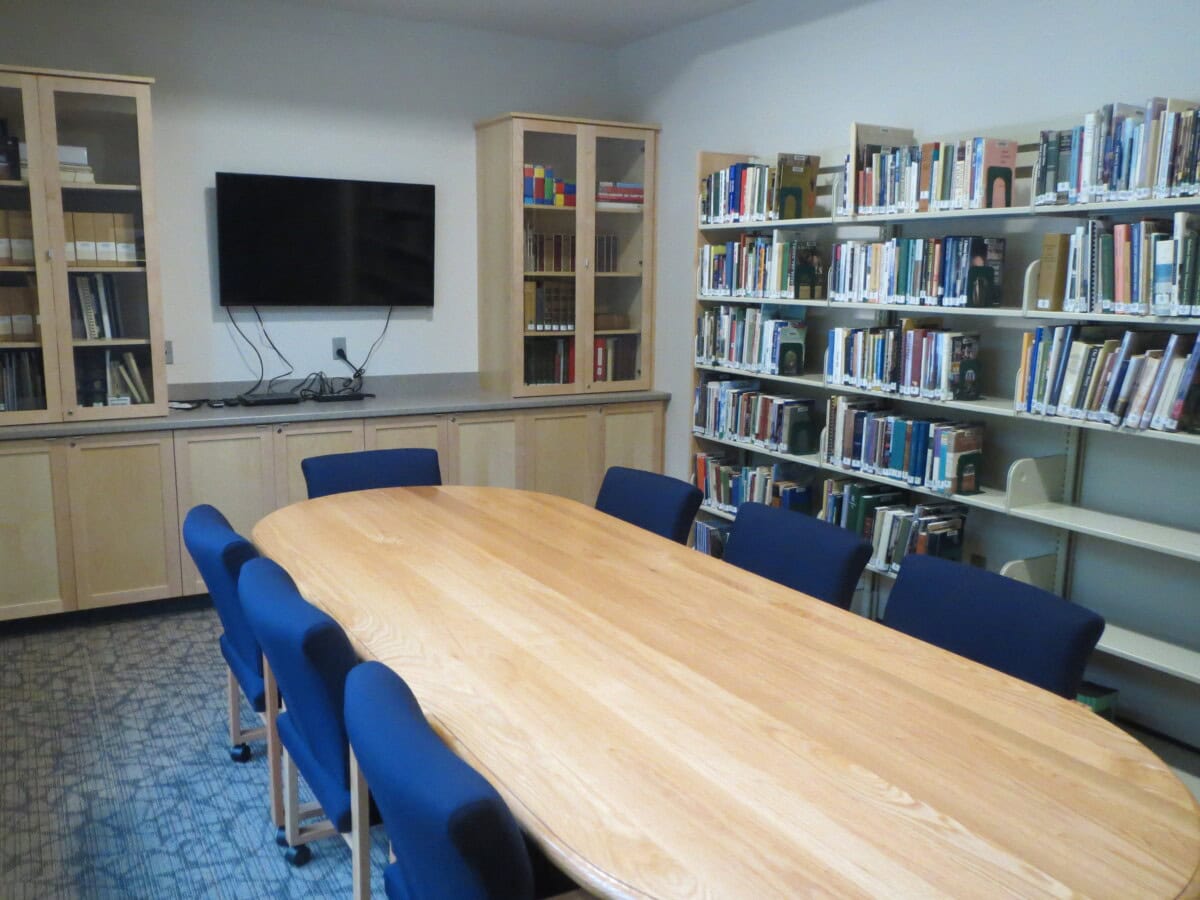 A conference table with blue chairs is surrounded by bookshelves and cabinets; a television is mounted on the wall in a small library or meeting room.