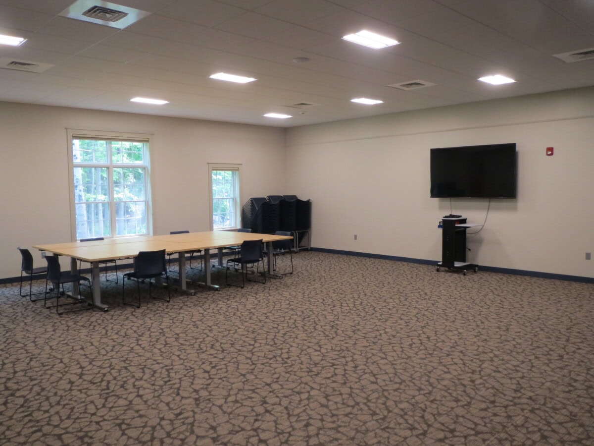 A meeting room with a rectangular table surrounded by chairs, a stack of extra chairs in the corner, large windows, and a wall-mounted TV with a stand below it.