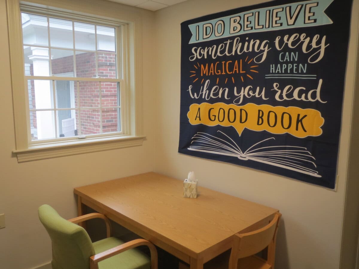 A small study nook with a window, wooden desk, two chairs, tissues, and a wall tapestry with a reading quote.